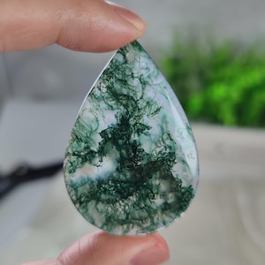 May include: A teardrop-shaped moss agate pendant featuring a translucent white base with intricate, dark green, fern-like dendritic inclusions throughout the stone. The polished gemstone has a small drilled hole at the top for suspension.