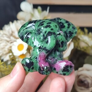 May include: A carved stone elephant figurine featuring a polished surface with a mottled pattern of vibrant green, deep black, and patches of bright magenta. The elephant is depicted in a seated position with its trunk curled toward its chest.