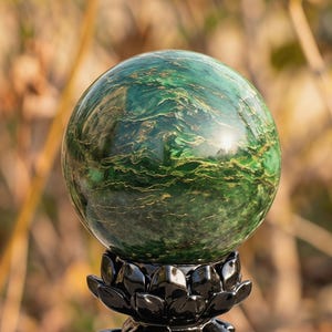 May include: A polished green sphere with gold veining sits atop a black lotus flower base. The sphere's surface reflects light, showcasing its intricate patterns. The background is blurred, emphasizing the sphere's details.
