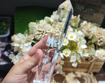 May include: A clear, six-sided quartz crystal point with a pointed tip and a flat base. The crystal features internal white inclusions and a smooth, polished surface that reflects light, showing a prismatic rainbow effect near the top apex.