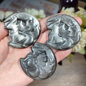 May include: Three circular silver obsidian carvings, each featuring a detailed relief of a nine-tailed fox curled within a crescent moon shape. The stones have a metallic, reflective sheen with smooth, polished surfaces and intricate engravings.