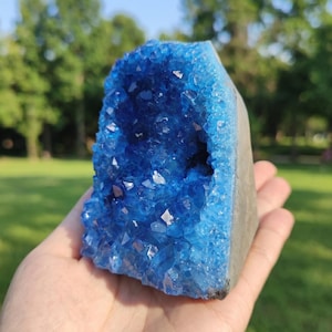 May include: A vibrant blue crystal geode with a rough, gray exterior. The interior is filled with sparkling, light blue crystal formations. The geode is held in a hand, with a blurred green background.