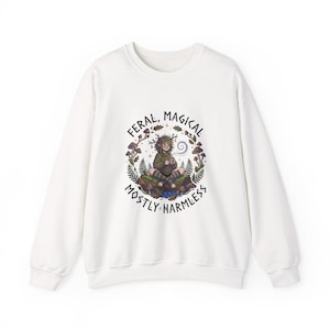 Könnte beinhalten: Weißes Sweatshirt mit einer Grafik, die eine sitzende Figur umgeben von Pilzen und Blättern zeigt. Der Text lautet "FERAL, MAGICAL, MOSTLY HARMLESS" in einem kreisförmigen Design. Die Illustration ist in Grün-, Braun- und Lilatönen gehalten.