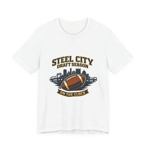 Puede incluir: Camiseta blanca con un diseño de fútbol americano. El texto "STEEL CITY DRAFT SEASON" está sobre un balón de fútbol y un horizonte urbano. El texto "ON THE CLOCK" está debajo.