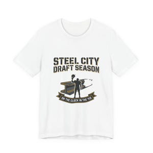 Peut inclure: T-shirt blanc avec l'inscription "STEEL CITY DRAFT SEASON" en noir et or. Le t-shirt présente un graphisme d'un joueur de football, d'un ballon de football et d'une poutre en I. Le texte "ON THE CLOCK IN THE 412" est également inclus.