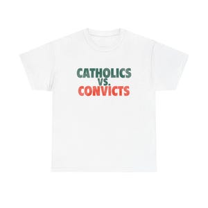 May include: White t-shirt featuring the text "CATHOLICS VS. CONVICTS" in green and red. The crew neck t-shirt is a casual style. The text is centrally positioned on the chest.