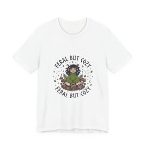 May include: White t-shirt featuring a whimsical illustration with the text "FERAL BUT COZY." The design includes a seated figure in a green sweater surrounded by mushrooms, stars, and crescent moons. The overall aesthetic is a blend of nature and fantasy.