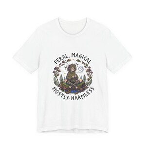 Könnte beinhalten: Weißes T-Shirt mit einer fantasievollen Illustration. Das Design zeigt eine sitzende Person inmitten von Blättern und Pilzen, mit dem Text "FERAL, MAGICAL, MOSTLY HARMLESS" in einem Kreis. Ein lässiges Kleidungsstück.