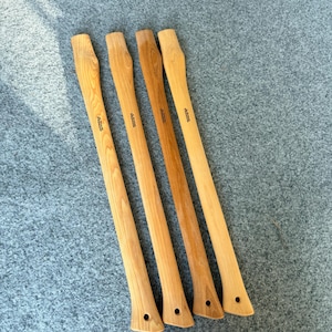 May include: Four long, slender axe handles crafted from light-coloured wood with visible grain patterns. Each handle features a curved shape, a flared end with a small circular hole, and a small brand logo stamped near the top section of the wood.