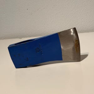 May include: A blue painted metal axe head featuring a polished steel blade edge. The axe head has a rectangular eye for a handle and a stamped circular logo on the side. The metal surface shows minor signs of wear and light oxidation near the edge.