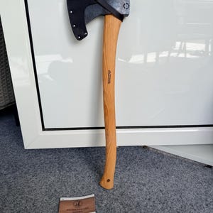 May include: A steel axe with a curved wooden handle and a black leather sheath covering the blade. The handle features a stamped logo and text reading Wetterlings Axes Sweden. A small rectangular product tag is included with the same branding.