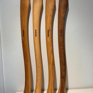May include: Four wooden axe handles arranged vertically, each featuring a distinct wood grain pattern and colour ranging from light tan to dark brown. Each handle is stamped with the text "Wetterlings" and "Made in Sweden" in black ink.