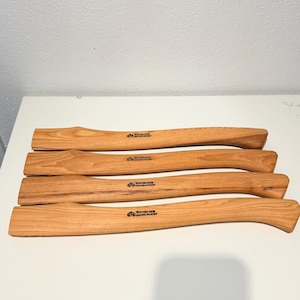 May include: Four light brown wooden axe handles arranged horizontally. Each handle features a curved shape and a smooth grain finish. The text Wetterlings Genuine Hickory is printed in black ink on the center of each handle, along with a small logo.