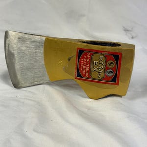 Könnte beinhalten: Ein Axtkopf aus Metall mit einer polierten silberfarbenen Klinge und einem goldfarbenen Körper. Auf der Seite befindet sich ein rechteckiges rot-weiß-blaues Etikett mit dem Text EXYXAEX EX und S.A. WETTERLINGS YXFABRIK STORVIK sowie kreisförmigen Logos.