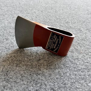 Könnte beinhalten: Ein Axtkopf aus Metall mit einer silberfarbenen Klinge und einem rot lackierten Körper. Auf der Seite befindet sich ein schwarzer, rechteckiger Aufkleber mit weißem Text: BUSQUESE LA ESTAMPA S.A. WETTERLING STORVIK SUECIA SOBRE CADA INSTRUMENTO.