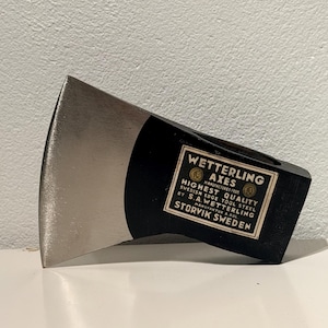 Może przedstawiać: Głowica siekiery z czarnego metalu z polerowanym srebrnym ostrzem. Z boku znajduje się prostokątna biała etykieta z napisem: WETTERLING AXES, HIGHEST QUALITY, SWEDISH EDGE TOOL STEEL, BY S.A. WETTERLING, MANUFAKTUR A.BOL, STORVIK SWEDEN.
