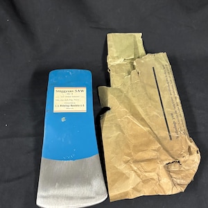 May include: A blue and silver axe head with the text "Släggyxan SAW" and a paper wrapping. The axe head has a blue top and a silver blade. The paper wrapping is brown and torn.