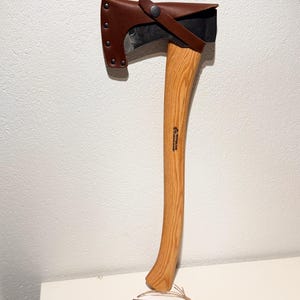 May include: A Wetterlings Axes Sweden hatchet with a curved light brown wooden handle and a dark metal head protected by a brown leather sheath with metal rivets and a snap closure. A rectangular tag with the text Wetterlings Axes Sweden is attached.