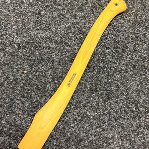 Op de afbeelding: Een gebogen bijlsteel van geel hickoryhout met een gladde afwerking en een klein gat aan de bovenkant. De steel is voorzien van een zwart gestempeld logo en de tekst Wetterlings Genuine Hickory, geplaatst in het midden van de houtnerf.