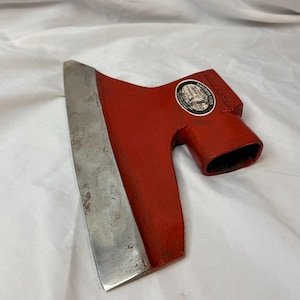 Puede incluir: Una cabeza de hacha pintada de rojo con un filo de metal plateado. La cabeza del hacha presenta una pegatina blanca circular con texto negro que dice SWEDISH AXES e incluye una ilustración gráfica de un árbol y una persona.