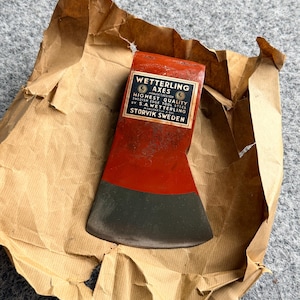 Puede incluir: Una cabeza de hacha de metal rojo con un filo gris oscuro, que presenta una etiqueta en blanco y negro que dice Wetterling Axes, Highest Quality Swedish Edge Tool Steel by S.A. Wetterling, Storvik Sweden, sobre papel marrón.
