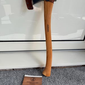 May include: A wood-handled axe with a dark metal head and a brown leather sheath. The axe has a curved handle and a sharp blade. The text "WETTERLINGS AXES SWEDEN" is visible on a small tag.