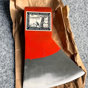May include: A bright red axe head with a polished silver metal blade edge. The axe head features a white rectangular label with black text reading MACHADOS SUECOS, O-RACHADOR, S.A. WETTERLING, and FABRICADOS NA SUÉCIA, along with a graphic illustration.