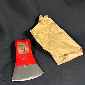 Op de afbeelding: Een rood en grijze bijlkop met een papieren verpakking. De bijlkop heeft een rood geverfde bovenkant en een grijs blad. De bijlkop heeft tekst en een afbeelding op het rode gedeelte. De papieren verpakking is bruin.