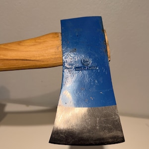 Puede incluir: Un hacha con una cabeza de metal pintada de azul y un mango de madera de color marrón claro. La cabeza del hacha presenta un sello circular con un logotipo de corona y el texto "MADE IN SWEDEN" grabado en el lateral. El filo es de metal plateado.