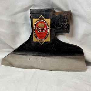Könnte beinhalten: Ein schwarzer Axtkopf aus Metall mit einer polierten silbernen Schneide. Auf der Seite befindet sich ein rautenförmiges gelbes Etikett mit dem Text S.A. WETTERLINGS YXFABRIK STORVIKS YXAN STALAD MED T-HA KROMSTAL FABR. GR 1880 und zwei Goldmünzen.