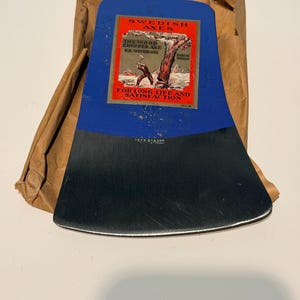 Könnte beinhalten: Ein blau-silberner Axtkopf aus Metall mit einem rechteckigen Etikett, das den Text SWEDISH AXES, THE WOOD CHOPPER AXE, S.A. WETTERLING, STORVIK SWEDEN und FOR LONG LIFE AND SATISFACTION enthält. Die Metalloberfläche ist mit DROP FORGED geprägt.
