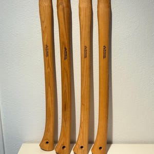 Könnte beinhalten: Vier hellbraune Axtstiele aus Holz, die vertikal angeordnet sind. Jeder Stiel hat eine geschwungene Form, ein verbreitertes Ende mit einem kreisförmigen Loch und den schwarzen Aufdruck Wetterlings Genuine Hickory im oberen Bereich des Holzes.