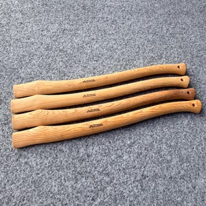 May include: Four light brown wooden axe handles arranged in a parallel row. Each handle features a curved shape, a smooth grain texture, and a small hole at the end. A small black logo with the text Wetterlings is printed on each handle.