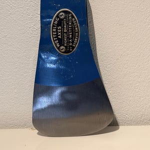 Op de afbeelding: Een blauwe metalen bijlkop met een gepolijste zilveren snijkant. Een ovale sticker op de zijkant vermeldt: Wetterling Axes, Highest Quality, Manufactured from Swedish Edge Tool Steel by S.A. Wetterling, Manufaktur A. Bol., Storvik Sweden.