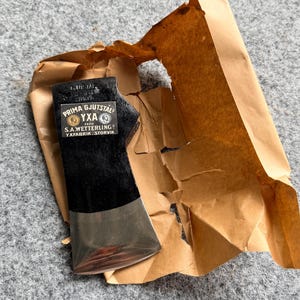 May include: A black metal axe head with a polished silver edge, partially wrapped in torn brown paper. The axe head features a prominent label with the text: PRIMA GJUTSTÅL YXA FRAN S.A.WETTERLING YXFABRIK STORVIK, including two circular emblem icons.