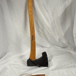 Könnte beinhalten: Eine Axt mit langem Holzstiel mit geschwungener Maserung und einem dunklen Metallkopf, der durch eine schwarze Lederscheide geschützt ist. Der Stiel hat ein kleines Loch am Ende. Ein rechteckiges Etikett mit dem Text Wetterlings Axes Sweden liegt bei.