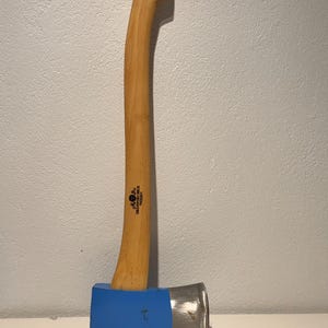 May include: A felling axe with a long, curved light-coloured wooden handle and a blue-painted metal head. The handle features a circular hole at the top and the text Gränsfors Bruk Sweden printed in black ink near the base of the axe head.