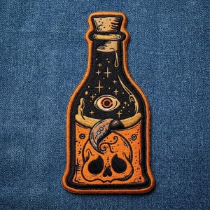 May include: An embroidered patch shaped like a bottle with a cork stopper. The design features a black upper section with a central eye and stars, and an orange lower section containing a skull and a curved horn, all outlined in orange thread.