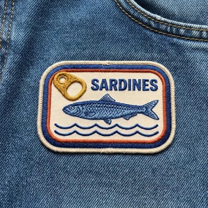 May include: A rectangular embroidered patch attached to blue denim fabric. The patch features a cream background with a blue border, an orange inner line, and the word SARDINES in blue. It depicts a blue fish, waves, and a gold pull tab.