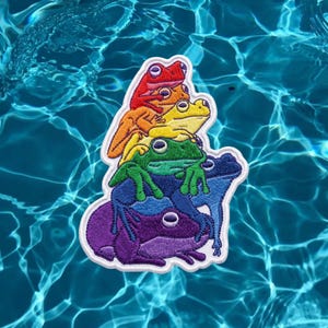 May include: An embroidered patch featuring a vertical stack of five frogs in a rainbow color sequence. The frogs are colored red, orange, yellow, green, blue, and purple from top to bottom, all outlined in white thread against a water background.