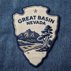 May include: A shield-shaped embroidered patch featuring a cream-colored background with a dark blue border. The design depicts a mountain range and a tree in dark blue and tan, with the text GREAT BASIN NEVADA, a star, and the year 1986.