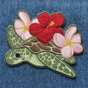 May include: An embroidered patch featuring a light green sea turtle with a brown shell pattern. The turtle is adorned with one large, vibrant red hibiscus flower and two smaller, pale pink plumeria flowers with yellow centers on its back.