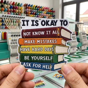 May include: A rectangular embroidered patch featuring a stack of five colorful books with the text IT IS OKAY TO NOT KNOW IT ALL, MAKE MISTAKES, HAVE HARD DAYS, BE YOURSELF, and ASK FOR HELP stitched in black and white thread on the book spines.