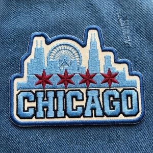May include: An embroidered patch featuring the Chicago skyline in light blue thread against a cream background. The design includes a Ferris wheel, four red six-pointed stars, and the word CHICAGO in bold, light blue block letters with dark blue outlines.