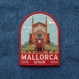 May include: An embroidered patch featuring a detailed depiction of a terracotta-colored cathedral against a light blue sky with white clouds. The base is a deep red border with the white text MALLORCA SPAIN and horizontal lines.