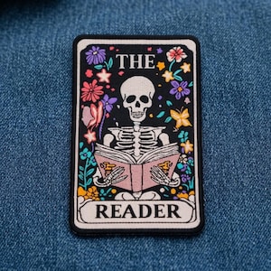 May include: A rectangular embroidered patch featuring a white skeleton reading a pink book. The design is surrounded by colorful flowers and butterflies on a black background. The text THE READER is printed in black within white borders.