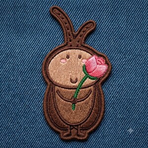 May include: An embroidered patch featuring a brown, cartoon-style insect with long antennae, a rounded body, and small feet. The insect has a light brown face with pink cheeks and is holding a single pink rose with a green stem in its arms.