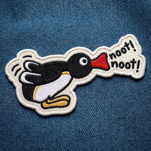 May include: An embroidered patch featuring a black and white penguin with a red beak, yellow feet, and a speech bubble containing the black text noot! noot! The patch has a cream-colored border and is positioned against a blue denim fabric background.