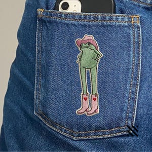 May include: An embroidered patch on a blue denim pocket featuring a green frog wearing a pink cowboy hat and matching pink cowboy boots with small red heart details. The frog stands with hands on hips, and a black smartphone is visible above.