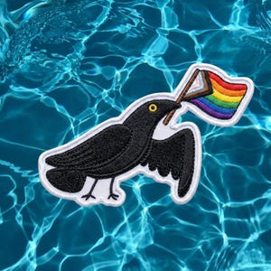 May include: An embroidered patch featuring a black crow with a yellow eye, holding a small flag in its beak. The flag displays the Progress Pride design, incorporating the traditional rainbow stripes alongside white, pink, blue, brown, and black colors.